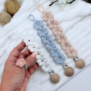 Op de afbeelding: Gehaakte fopspeenclips in wit, lichtblauw en perzik. Elke clip heeft een ketting van gehaakte bloemen en een houten clip. Een gehaakte bloemenring is ook zichtbaar. De items worden gepresenteerd op een wit, getextureerd oppervlak.