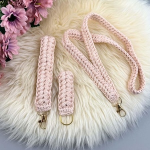 May include: A set of four light pink crocheted accessories: a lanyard, two keychains, and a wristlet. Each item features a braided design and gold-colored hardware. The items are displayed on a fluffy, cream-colored surface, with pink flowers in the background.