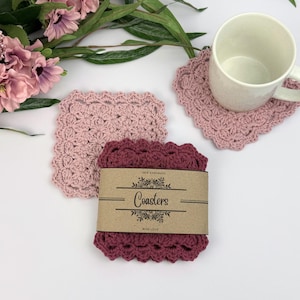May include: Handmade crochet coasters in pink and burgundy. A white mug sits on a pink coaster. A stack of coasters is wrapped with a brown paper band that reads "Coasters" and "With Love".