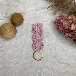 May include: A pink crocheted keychain with a gold metal ring. The keychain is made of a delicate, floral pattern.