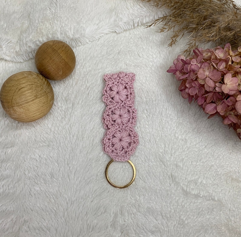 May include: A pink crocheted keychain with a gold metal ring. The keychain is made of a delicate, floral pattern.