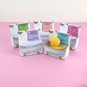 Puede incluir: Cinco peque&ntilde;as cajas de regalo con tem&aacute;tica de ba&ntilde;o con patitos de goma amarillos en su interior. Cada caja tiene un mensaje diferente en la parte delantera, incluyendo "Si la vida te da limones, s&eacute; un pato", "En un mundo lleno de palomas, s&eacute; un pato fabuloso", "&iexcl;Me haces sentir tan pato!" y "&iexcl;Te amo pato!"
