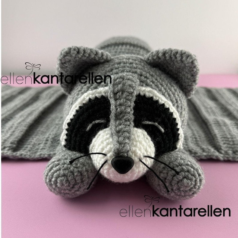 May include: A handmade crochet raccoon blanket in shades of gray, black, and white. The raccoon has a black mask, white face, and a black nose. The blanket is rectangular with a ribbed texture. The text "ellenkantarellen" is visible.
