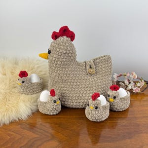 May include: A crocheted chicken-shaped basket with a red comb and yellow beak, accompanied by four smaller crocheted chicken egg holders. Each holder has a red comb and holds a white egg. The items are beige.