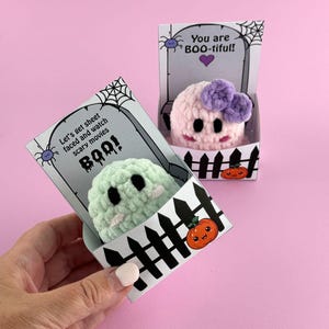 Puede incluir: Dos peluches de fantasmas de ganchillo en cajas decorativas. Uno es verde claro con ojos negros y una cara blanca, el otro es rosa con un lazo morado. Las cajas tienen un dise&ntilde;o de l&aacute;pida con el texto "Let's get sheet faced and watch scary movies BOO!" y "You are BOO-tiful!"