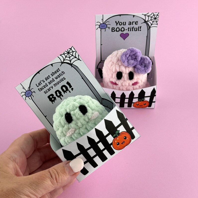 Puede incluir: Dos peluches de fantasmas de ganchillo en cajas decorativas. Uno es verde claro con ojos negros y una cara blanca, el otro es rosa con un lazo morado. Las cajas tienen un dise&ntilde;o de l&aacute;pida con el texto "Let's get sheet faced and watch scary movies BOO!" y "You are BOO-tiful!"