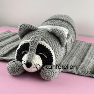 May include: A gray crocheted raccoon-shaped cozy with a white and black face, black nose, and whiskers. The cozy has a striped tail and is designed to hold a cup or mug. The text "Elle kantarellen" is visible on the bottom right.
