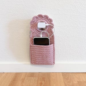 Könnte beinhalten: Ein wandmontierter Handyhalter aus altrosa Häkelarbeit mit einem gewellten Oberteil, einer Tasche und einem weißen Ladegerät, das in eine Steckdose gesteckt ist. Ein schwarzes Smartphone befindet sich in der Tasche. Der Halter ist an einer weißen Wand befestigt.