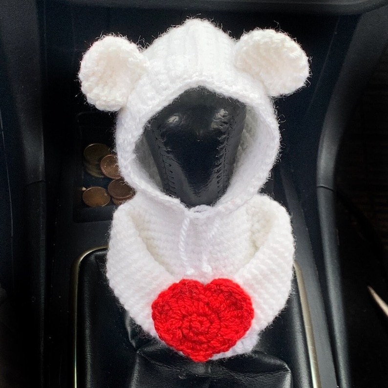 May include: White crocheted stuffed animal wearing a hooded sweater with a red heart. The animal has two small ears and is sitting in a car.