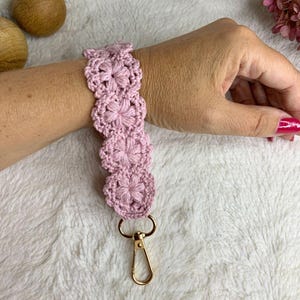 May include: A light pink crocheted wristlet featuring a floral design. The wristlet is made of soft yarn and has a gold-colored clasp. The floral pattern is repeated along the length of the wristlet, creating a delicate and feminine accessory.