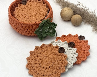 May include: A set of four crocheted coasters in orange and white, with a green crocheted leaf, in a woven orange basket. The coasters are round and have a raised, textured design.