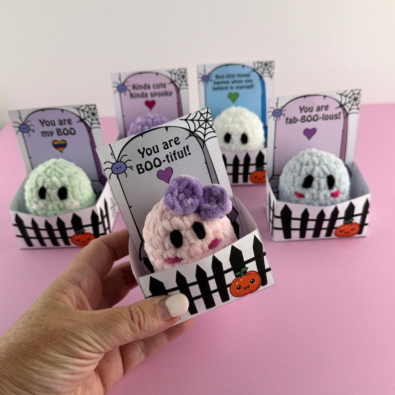 Puede incluir: Una colecci&oacute;n de peluches de fantasmas hechos a mano con ganchillo en varios colores pastel, cada uno dentro de una caja decorativa con forma de valla. Las cajas presentan texto y gr&aacute;ficos con tem&aacute;tica de Halloween, incluyendo frases como "You are BOO-tiful!" y peque&ntilde;as calabazas.