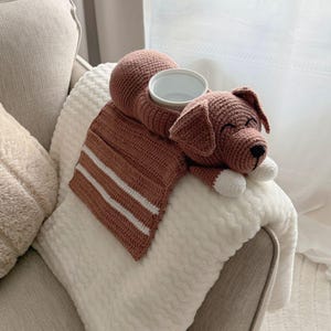 K&ouml;nnte beinhalten: Ein handgefertigter, geh&auml;kelter Hunde-Becherhalter in warmem Braun, mit wei&szlig;en Akzenten an den Pfoten und einer wei&szlig;en Schale. Der Hund liegt auf einer wei&szlig;en Decke mit einem braun-wei&szlig; gestreiften Schal.