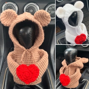 May include: Three crocheted car gear shift covers in the shape of hooded animals. The covers are brown, white, and brown with red hearts on the front.