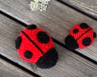 May include: Two crocheted ladybugs, one red with black spots and a black head, the other smaller and red with black spots and a black head, on a wooden surface.
