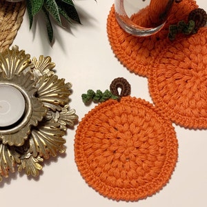 May include: Orange crochet pumpkin coasters with green and brown accents. The coasters are round and have a textured appearance. A gold candle holder and a small plant are also visible.