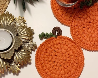 May include: Orange crochet pumpkin coasters with green and brown detailing. The coasters are circular and have a textured look. A gold candle holder and a small plant are also visible.