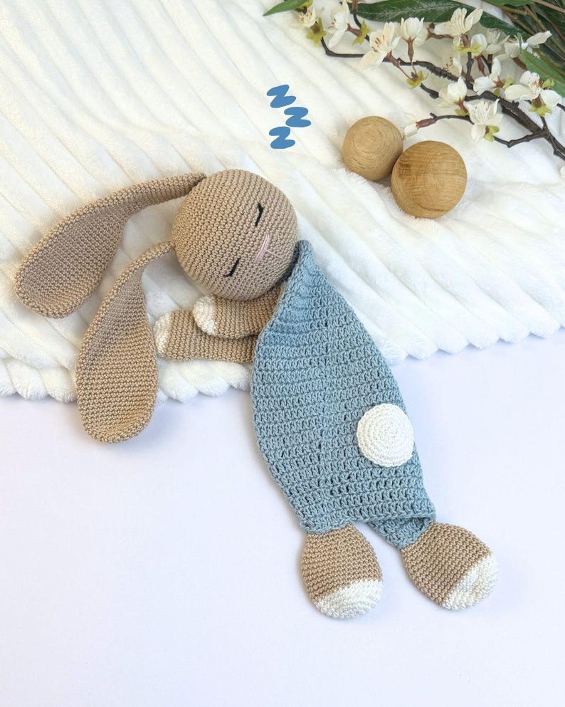 May include: A handmade crochet bunny toy in beige and light blue. The bunny has long ears, closed eyes, and a blue blanket. The bunny's feet are beige with white tips. Two wooden spheres and white flowers are in the background.
