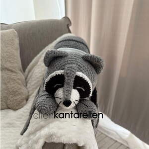 May include: A handmade crochet raccoon in gray, black, and white, resting on a white plush blanket. The raccoon has a black mask, white face, and a striped tail. The text "ellenkantarellen" is visible.
