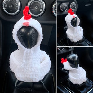 Könnte beinhalten: Weißer, gehäkelter, huhnförmiger Automatik-Schalthebelbezug mit roten Akzenten. Der Bezug ist auf einem schwarzen Schalthebel in einem Auto angebracht.