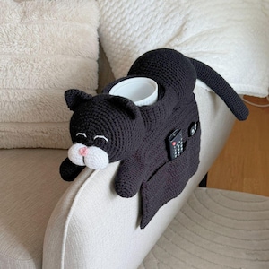 May include: A black crocheted cat-shaped couch armrest organizer. The cat has a white muzzle and pink nose. It features a cup holder and pockets for a remote control and other small items. The cat is lying on a beige couch arm.