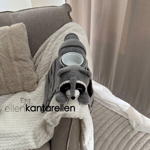 May include: A gray and white crocheted raccoon-shaped armrest cup holder. The raccoon has black and white facial features and is holding a white ceramic bowl. The product is placed on a gray sofa arm.