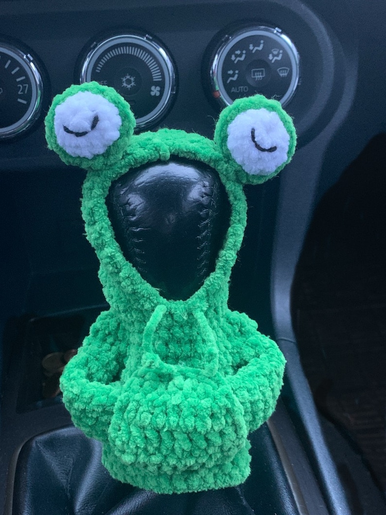 Pu&ograve; includere: Copri leva del cambio in crochet verde a forma di rana con occhi bianchi e bocca nera. La copertura &egrave; sulla leva del cambio di un'auto.