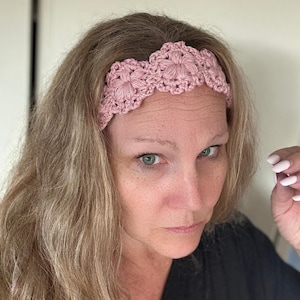 May include: A dusty pink crochet headband featuring a repeating floral pattern with scalloped edges. The accessory is constructed from yarn with a textured, dimensional stitch design that creates a series of connected circular motifs across the band.