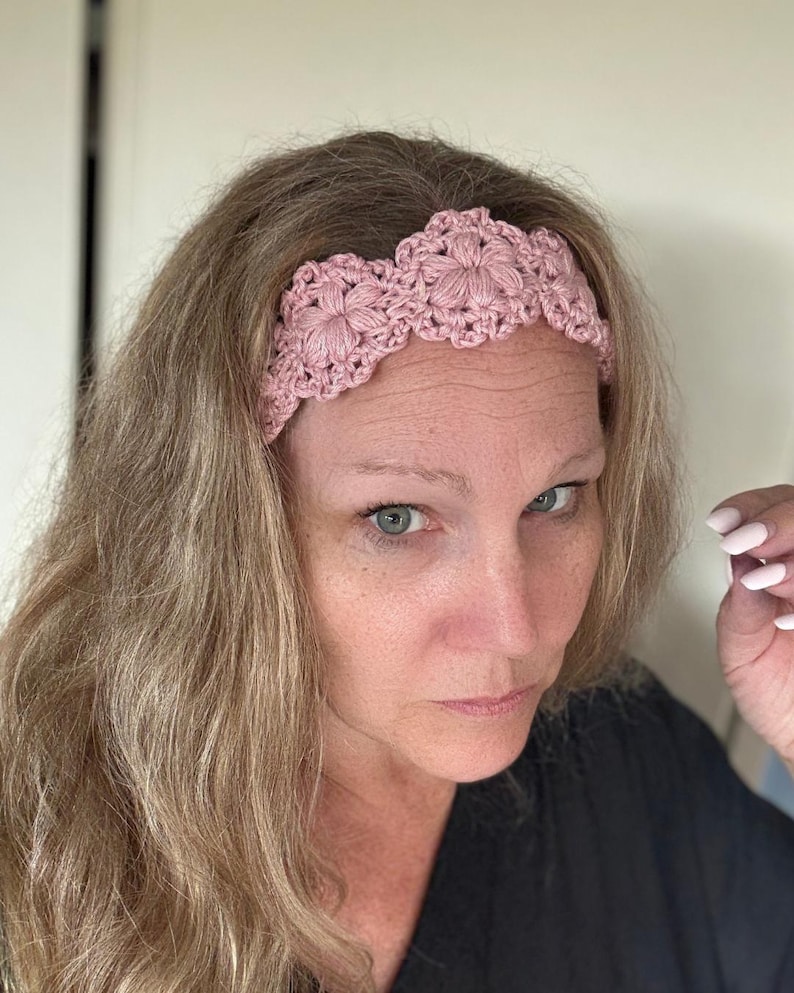 May include: A dusty pink crochet headband featuring a repeating floral pattern with scalloped edges. The accessory is constructed from yarn with a textured, dimensional stitch design that creates a series of connected circular motifs across the band.