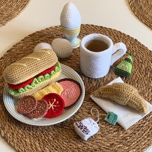 May include: Crocheted breakfast food set with a sandwich, egg, croissant, tea bag, and mug. The set is on a brown woven placemat.