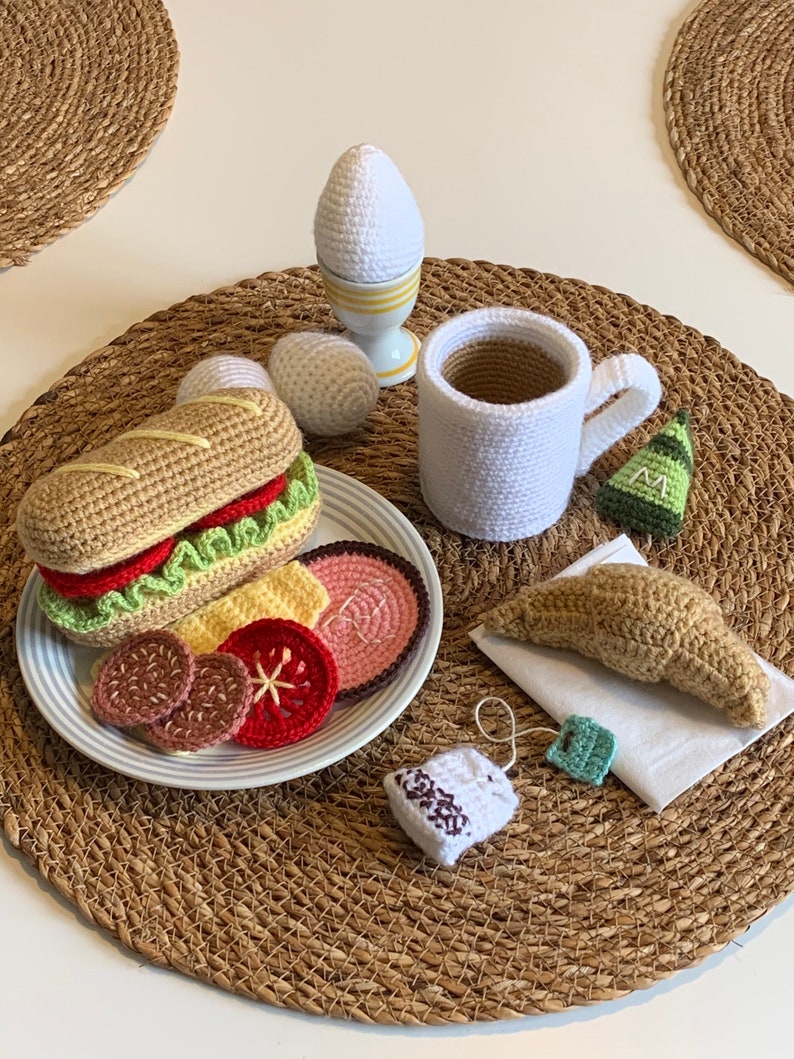 May include: Crocheted breakfast food set with a sandwich, egg, croissant, tea bag, and mug. The set is on a brown woven placemat.