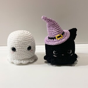 Puede incluir: Dos figuras de Halloween tejidas a crochet: un fantasma blanco y un gato negro con un sombrero de bruja morado. El fantasma tiene un dise&ntilde;o sencillo con mejillas rosadas. El gato tiene ojos grandes y un sombrero con una banda negra y hebilla amarilla. Ideal para decoraci&oacute;n de Halloween.