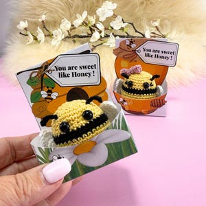 Puede incluir: Figuras de abeja de ganchillo en cajas decorativas. Las abejas son amarillas y negras, con antenas negras y ojos grandes negros. Las cajas presentan un diseño de panal y tarro de miel, con el texto "You are sweet like Honey!"