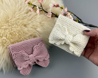 May include: Two crochet headbands, one light pink and one cream, each featuring a large, decorative bow. The bows have a scalloped edge. The headbands are displayed on a fluffy, cream-colored surface with pink flowers.