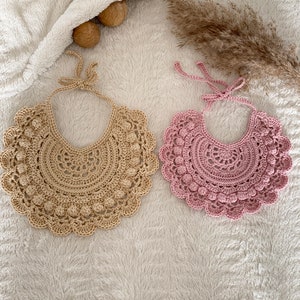 May include: Two crocheted collars, one beige and one pink, with a scalloped edge and a decorative bead pattern. The collars are laid out on a white fluffy surface.