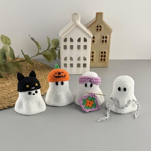 May include: Four white crochet ghost figures, each with a unique hat or accessory. One ghost wears a black cat hat, another a pumpkin hat, a third a purple headband, and the last has a crochet chain. Decorative Halloween theme.