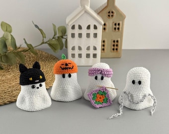 May include: Four white crochet ghost figures, each with a unique hat or accessory. One ghost wears a black cat hat, another a pumpkin hat, a third a purple headband, and the last has a crochet chain. Decorative Halloween theme.