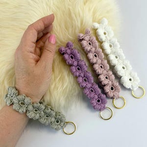 May include: A selection of handmade macrame flower keychains in shades of sage green, purple, pink, and white. Each keychain has crocheted flowers and a gold-coloured ring. A matching bracelet is also shown.