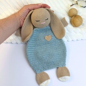 May include: A handmade crochet bunny in beige and light blue. The bunny has long floppy ears, closed eyes, and a small pink nose. It wears a light blue dress with a heart-shaped applique. The bunny's feet have white accents.