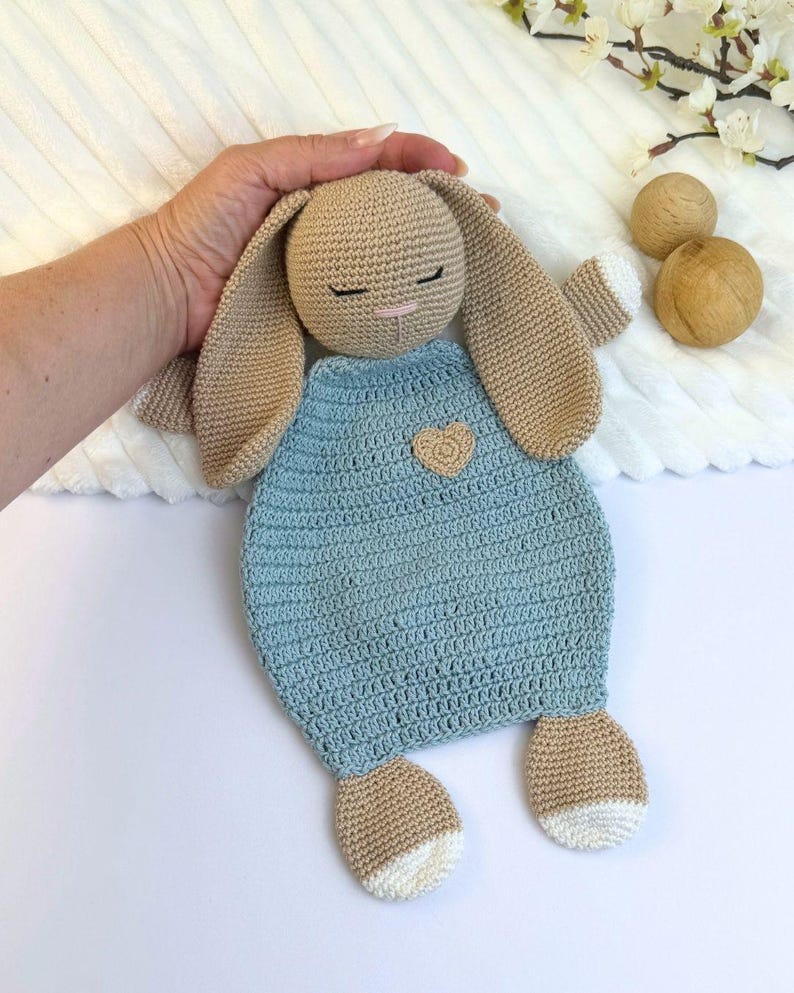 May include: A handmade crochet bunny in beige and light blue. The bunny has long floppy ears, closed eyes, and a small pink nose. It wears a light blue dress with a heart-shaped applique. The bunny's feet have white accents.