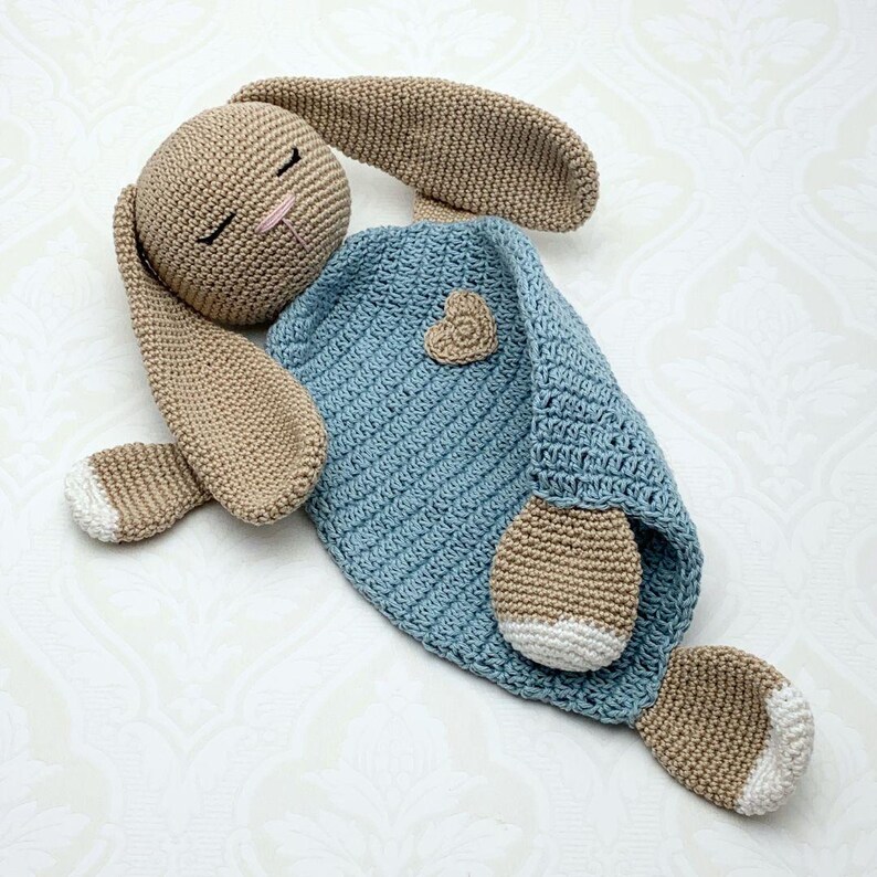 May include: A handmade crochet bunny security blanket in tan and light blue. The bunny has long ears, closed eyes, and a pink nose. The blanket is light blue with a small heart applique. The bunny's feet have white accents.