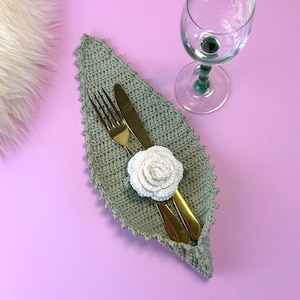 May include: A sage green crocheted leaf-shaped napkin with a white crocheted rose, holding a gold fork and knife. A clear wine glass with a green stem sits on a pink surface. The napkin adds a decorative touch to a dining table setting.