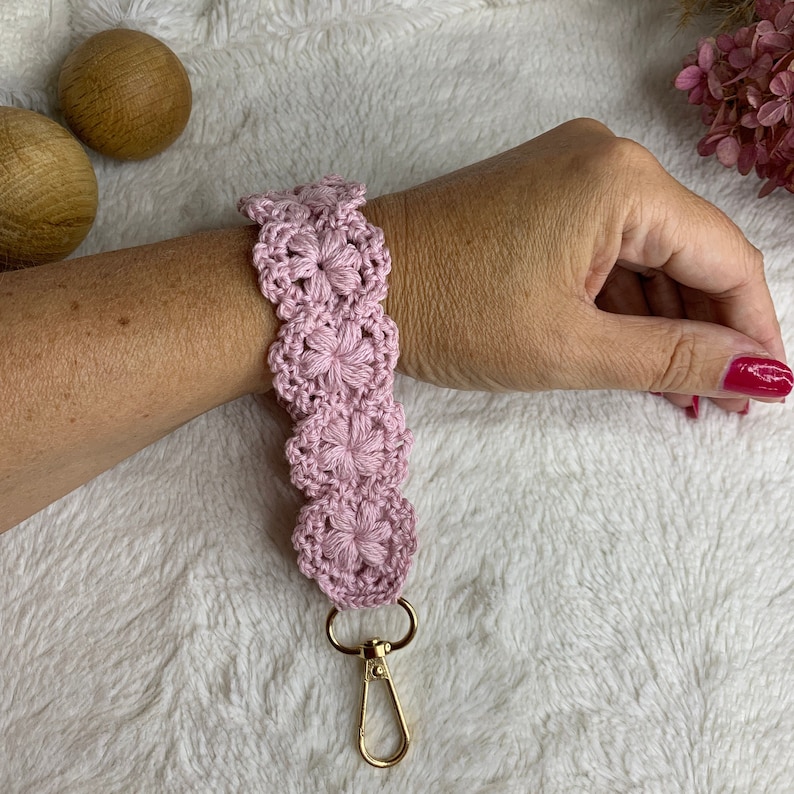 May include: A pink crocheted keychain with a gold metal clasp. The keychain is made of a series of crocheted flowers.