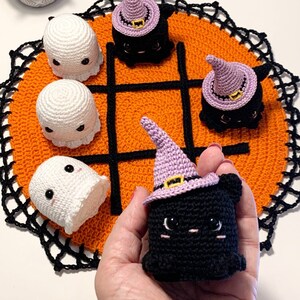 Puede incluir: Un juego de tic-tac-toe de ganchillo con tem&aacute;tica de Halloween. El tablero es naranja con l&iacute;neas negras y un borde de encaje negro. Las piezas del juego incluyen fantasmas blancos y gatos negros con sombreros de bruja morados.