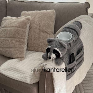 May include: A gray crocheted raccoon-shaped sofa arm organizer with a white bowl holder and two pockets. The raccoon has a white face, black eyes, and a striped tail. The organizer is placed on a sofa with beige pillows and a white blanket.