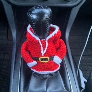 Puede incluir: Un disfraz de Pap&aacute; Noel de ganchillo para una palanca de cambios de coche. El disfraz es rojo y blanco con un cintur&oacute;n negro y un ribete blanco alrededor de la capucha.