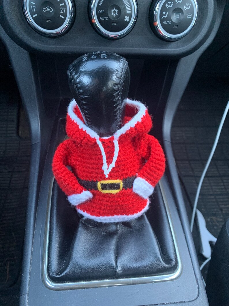 Puede incluir: Un disfraz de Pap&aacute; Noel de ganchillo para una palanca de cambios de coche. El disfraz es rojo y blanco con un cintur&oacute;n negro y un ribete blanco alrededor de la capucha.