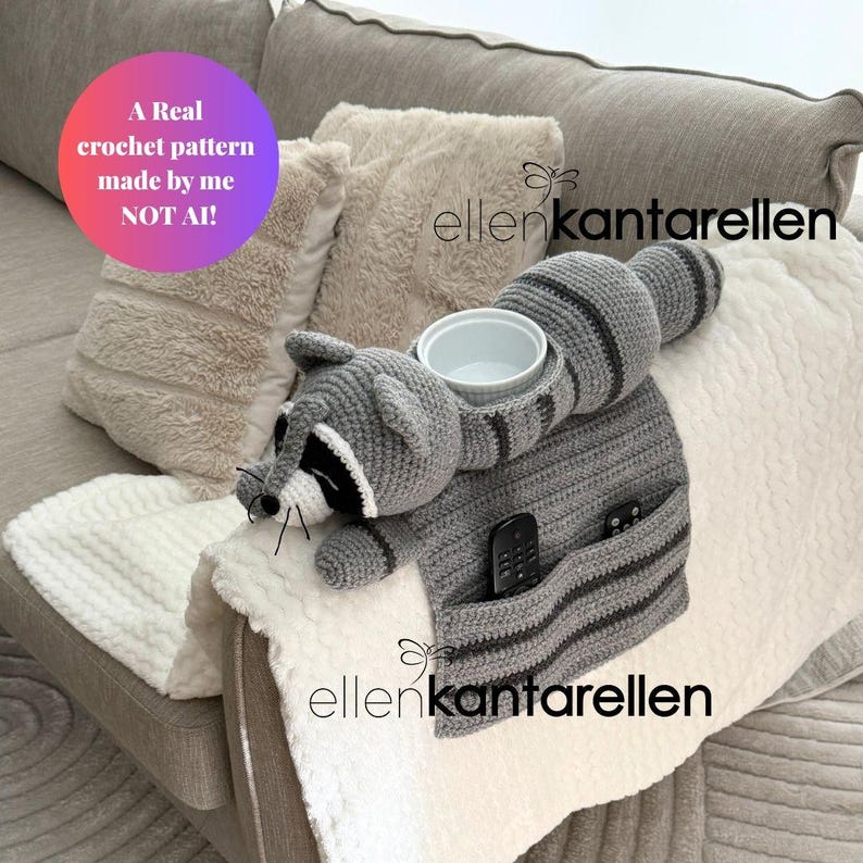 May include: A handmade crochet raccoon sofa caddy in gray and black, with a white bowl and pockets for a remote control. The caddy is placed on a white blanket over a sofa. Text on the image reads "A Real crochet pattern made by me NOT AI!"