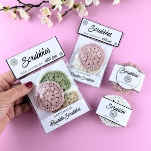 May include: Several handmade reusable scrubbies in various colors, including pink, green, and cream, are displayed on a pink background. Each scrubbie is packaged with a label that reads "Scrubbies" and "100% Handmade With Love."
