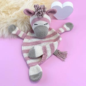 May include: A handmade crochet zebra comfort blanket in pink, white, and grey. The zebra has a striped body, a grey snout, and closed eyes. The blanket has a tail with a pink tassel. The background is a light pink surface.